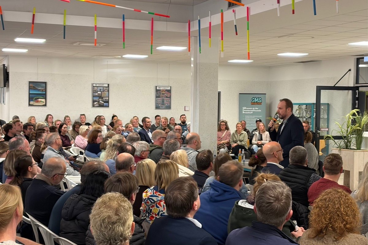 ein Mann steht vor vielen sitzenden Menschen und spricht