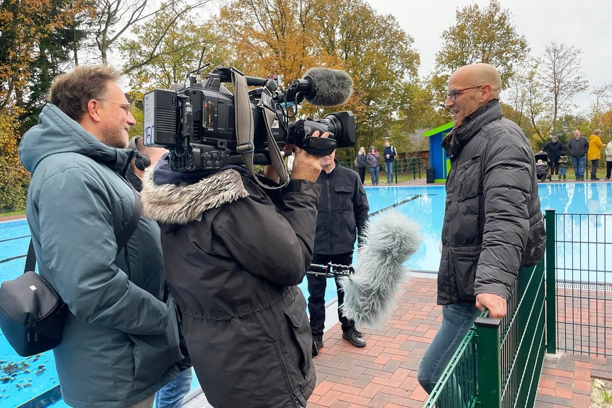 viel Männer stehen am Schwimmbadrand. einer hält eine Fernsehkamera und einer ein Mikrofon in Richtung eines anderen Mannes