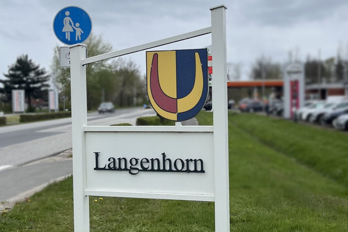 Langenhorn möchte mittels einer Umfrage Erkenntnisse über das Einzugsgebiet der Gemeinde erlangen. Foto: Oldsen/Langenhorn  Im Vordergrund sind ein Ortsschild sowie das Gemeindewappen zu sehen. Dahinter verläuft eine Straße