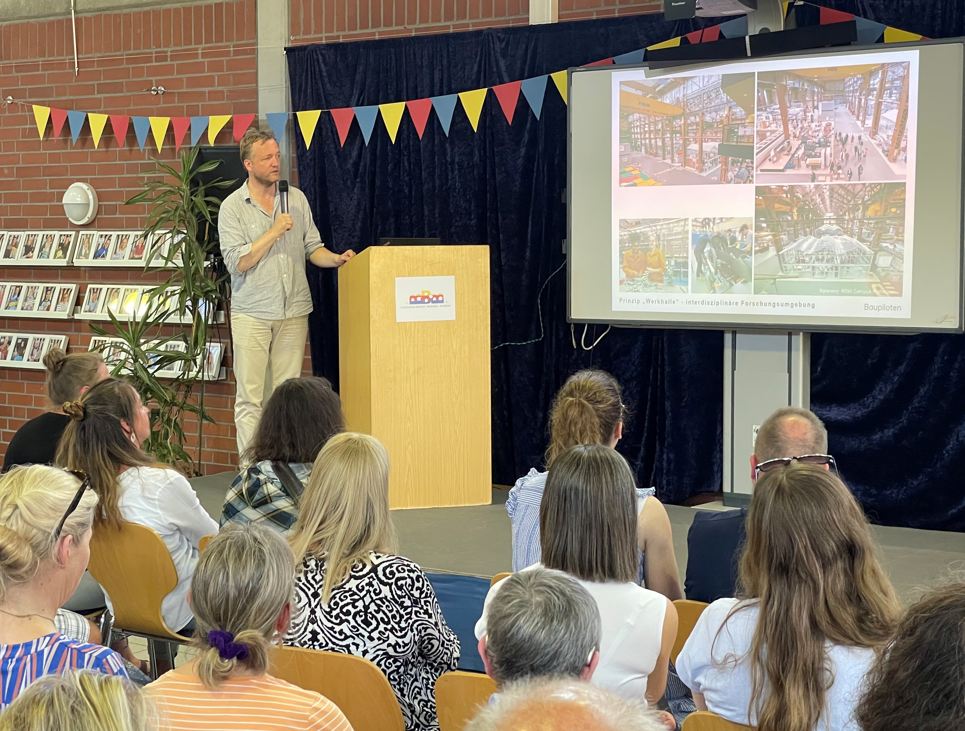 ein Mann steht auf einer Bühne und hält ein Mikrofon in der Hand. Vor ihm sitzt Publikum.